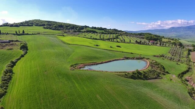 Aerial view of a small pond surrounded by lush green rolling hills and agricultural fields in the countryside of Tuscany, Italy.
