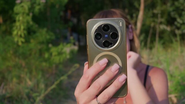 A woman holds a modern gold smartphone covering her face in a lush outdoor garden. The mobile phone features a prominent circular multi sensor array and protective magnetic case.
