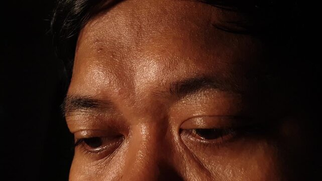 Close up frontal view of an adult Indonesian Asian man looking downward and slowly raising eyes upward in dark warm lighting, revealing forehead expression, skin texture, and serious