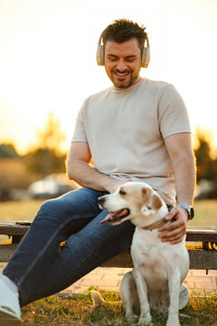 Man wearing headphones and petting his dog while relaxing on a bench at sunset
