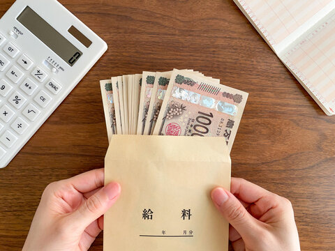 机の上で紙幣の入った給料袋を持つ女性の手／家計管理イメージ