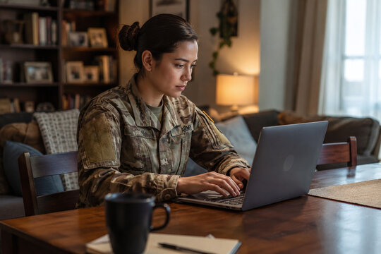Focused female soldier in camouflage uniform working on a laptop in a cozy home setting with a coffee cup. Concept of military mother, veteran education, and remote work lifestyle.