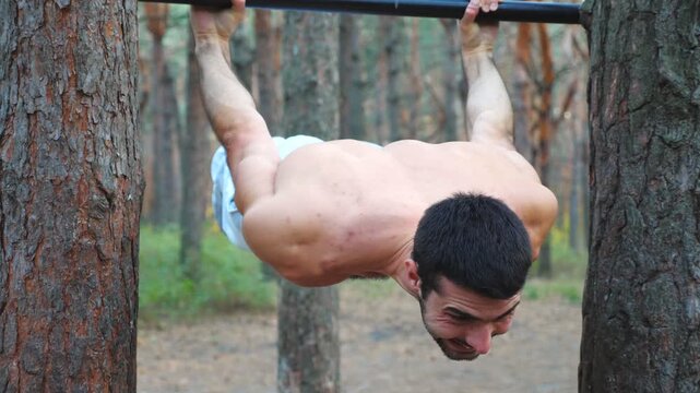 Strong man showing some gymnastics stunts on horizontal bar. Muscular guy doing gymnastics elements. Athlete performs strength exercises during workout in forest. Sportsman training static exercises