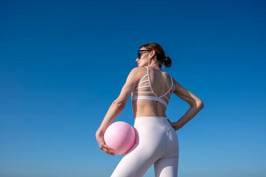 Fit woman in activewear holds a pink exercise ball outdoor, back view
