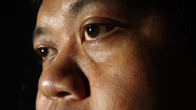 Extreme macro profile view of an adult Indonesian male left eye under warm golden directional light against a black background, featuring clear iris reflections and gentle eyelid m