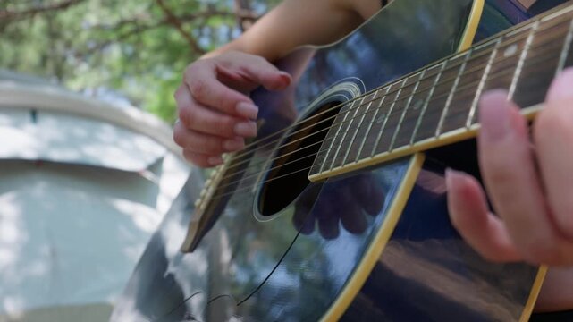 A songwriter hand plucks the steel strings of an acoustic guitar during a creative session outdoors. Natural daylight filters through green trees at a peaceful forest campsite.