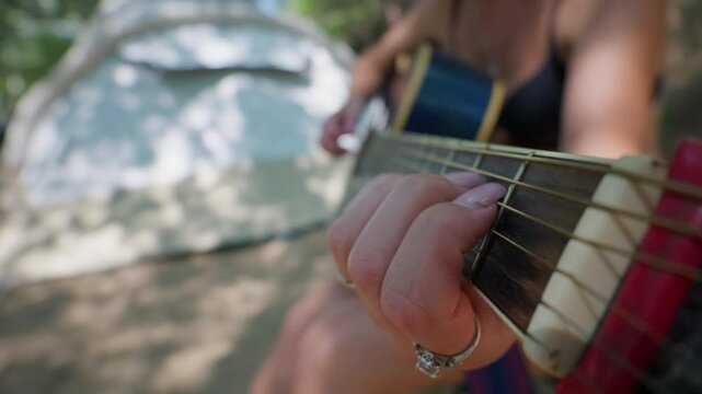 A close up of fingers pressing strings on an acoustic guitar fretboard while camping outside near a tent. Concept of musical recreation, acoustic melody, and outdoor summer leisure.