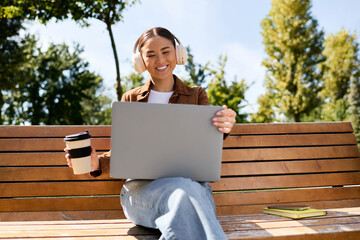 Fototapeta premium A cheerful young woman smiles on a bench, sipping coffee and using her laptop among trees.