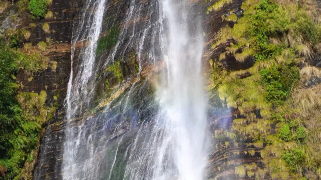 Rainbow Forms in Sunlit Waterfall Spray Over Mossy Mountain Rock Face