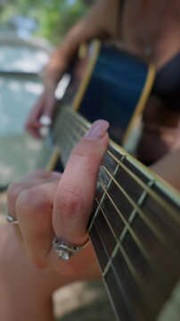 A woman fingers chord transitions on an acoustic guitar fretboard during outdoor music practice. Her manicured hand precisely presses the metal strings to learn a melody in daylight.