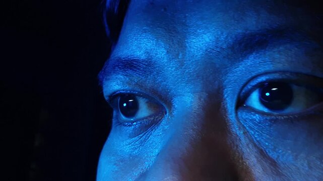 Extreme close up side view of an Indonesian male eyes looking sideways and reacting to changing red blue and green neon lights in a dark room highlighting detailed facial skin refl