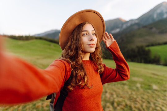 Selfie time! A woman hiker in a hat enjoys the sunset in a green meadow. A hiker with curly hair enjoys the mountain scenery and the freedom of the outdoors. Concept: blog, nature.