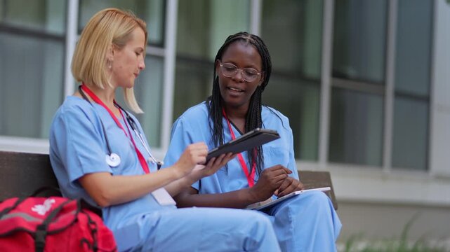 Two young female nurses in scrubs discussing a case using a digital tablet outside a hospital