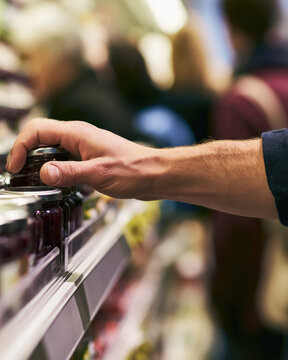 Crowded grocery store scene showing forearm and hand reaching for jar on clean metal shelf, with blurred shoppers, bright commercial lighting, realistic skin texture, and negative space on right