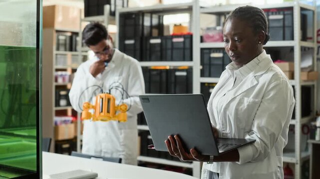 African American female engineer using laptop while Indian colleague inspects underwater robot in laboratory. Perfect for videos on robotics development and marine technology research