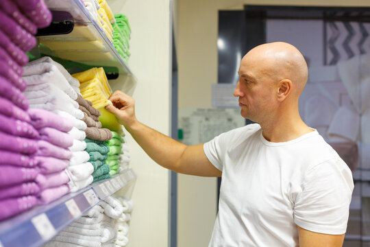 Bald caucasian man reaching for a folded towel on a shelf in a household goods shop, ideal for retail, consumer choice, shopping behavior and home textile content.