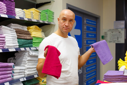 Mature caucasian man in a white shirt compares a pink and a purple towel at a shop shelf, weighing two options in the home textile department of a household goods store.