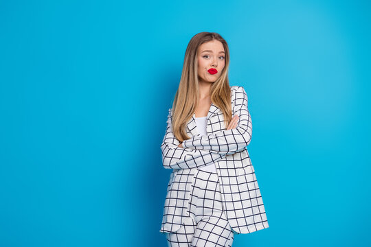 Confident young business woman in white checkered suit with crossed arms and playful pout on bright blue background