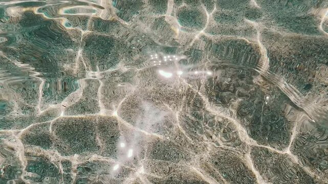 V&iacute;deo submarino art&iacute;stico de reflejos solares sobre arena bajo agua transparente.