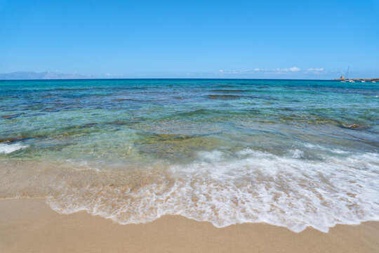 Clear turquoise Mediterranean Sea water washing onto a sandy beach on the coast of Sicily, Italy, with distant mountains visible on the horizon under a sunny summer sky.