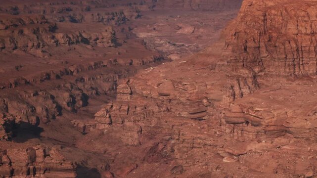 A group of people walks along the rocky terrain of a canyon area. The sun sets, casting warm light on the red rocks and creating long shadows. They enjoy the view and take pictures.