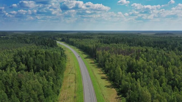 Aerial view of a single car driving on a long asphalt road cutting through a dense green forest under a blue sky with scattered clouds, Russia.