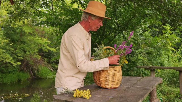A man collects various medicinal herbs near a river on a summer day. He uses a woven basket to hold the herbs while examining their properties closely