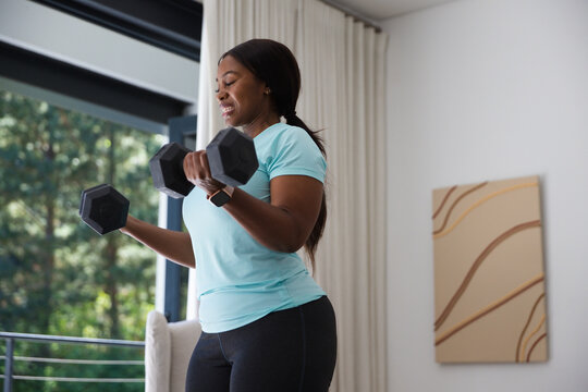 African American woman lifting hex dumbbells at home by glass door, wearing sportswear, smartwatch
