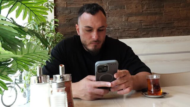 Focused young man sitting in a cafe using his smartphone and drinking tea