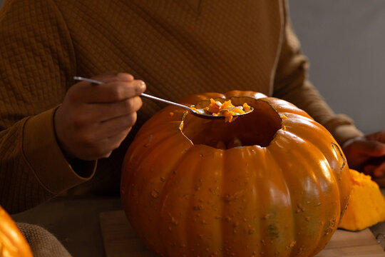 African American man scooping pumpkin pulp and seeds with spoon at home table wearing brown sweater