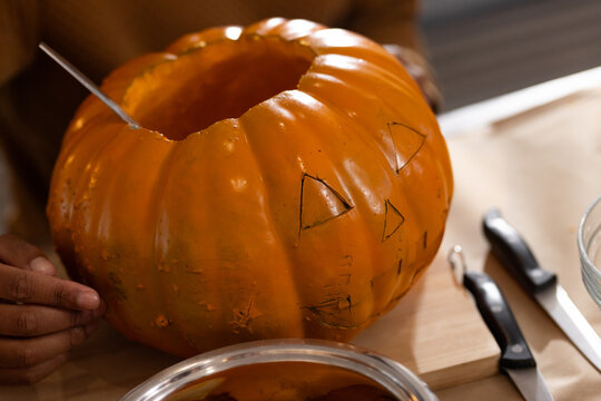 Hollow pumpkin is being scooped on wooden cutting board near knives and clear mixing bowl