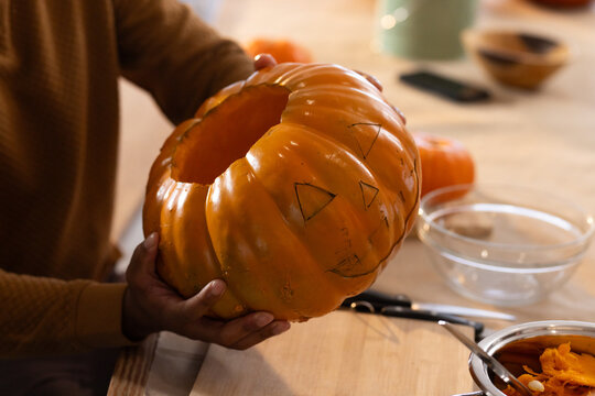 African American man in brown sweater holding hollowed pumpkin scooping pulp at wood table with pot