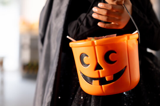 African American teenage female holding orange pumpkin candy bucket in hallway, wearing black cloak