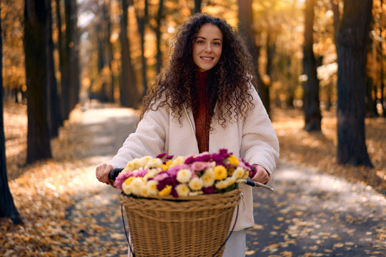 Portrait of a confident, smiling young beautiful woman standing with a bicycle featuring a wicker basket filled with flowers in an autumn park. Autumn recreation concept.