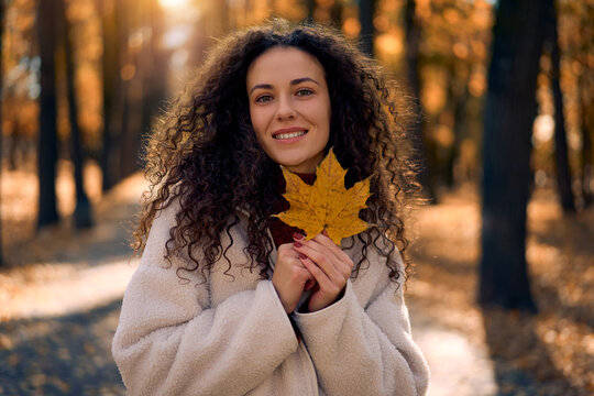 Portrait of a beautiful smiling young woman with curly hair holding a yellow maple leaf in a sunny autumn park.