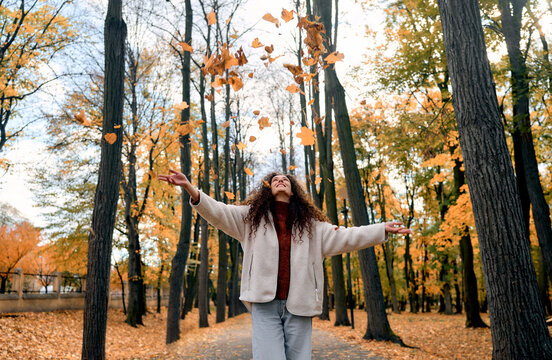 Young joyful woman looking up and enjoying falling yellow autumn leaves in a beautiful park. Happy carefree mood, seasonal nature, peaceful outdoor moment and autumn atmosphere.