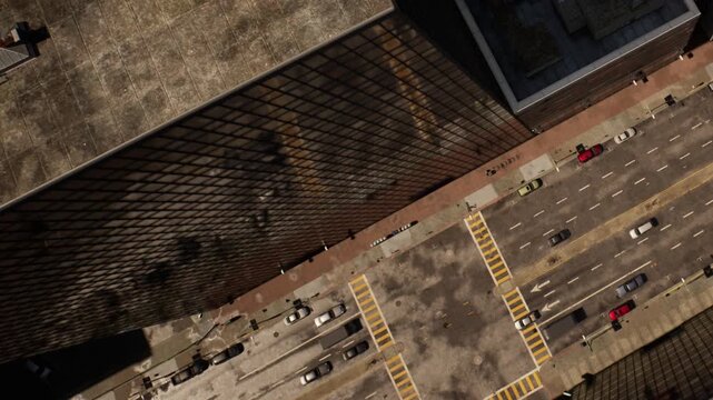 A view shows streets and parked cars below tall buildings in a city. The buildings shadows stretch across the ground, creating patterns. It is a clear day with no visible clouds.