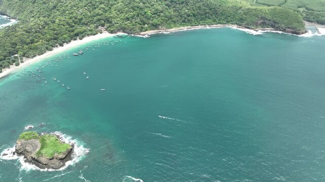 Aerial view of Papuma Beach with turquoise water, small fishing boats, and a rocky island off the lush green coastline in Jember, East Java, Indonesia.