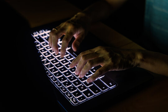 Close up of hands typing on illuminated backlit keyboard in dark room, cybersecurity hacker and programmer coding concept