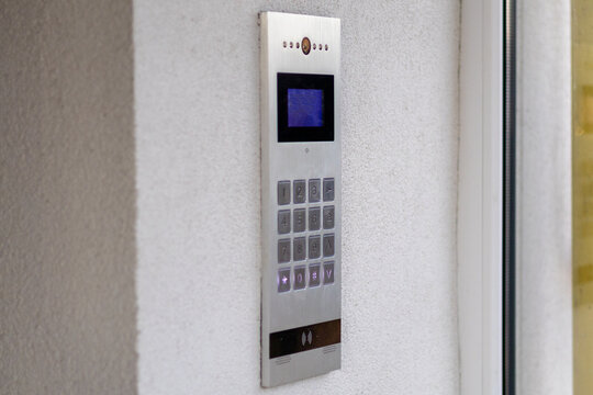 A silver intercom system is installed on a white wall next to a glass door outside a building. The device has buttons and a small screen. It is daylight and objects are clearly visible