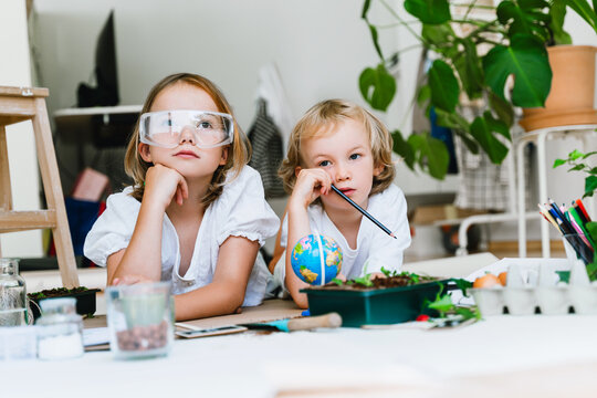 A little girl in safety glasses and a boy wearing white t-shirts lean on the table among plants. Creative home laboratory concept for ecology and futurism experiments.