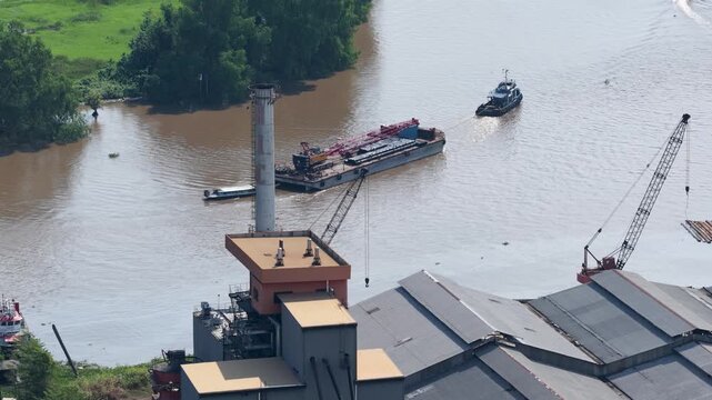 Heavy barge transport moving down the river past an industrial factory during the daylight hours