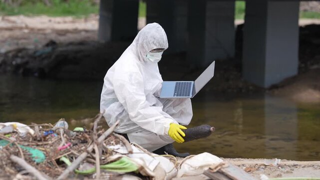 Environmental Scientist Protective Suit Uses Laptop for River Pollution Field Research, Environmental Monitoring, Water Quality Assessment, Water Pollution Study, Plastic Waste Documentation