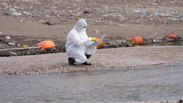 Environmental researcher protective suit and protective clothing with hood kneeling to collect water sample beside polluted shoreline, floating barrier, scattered waste, and polluted water during