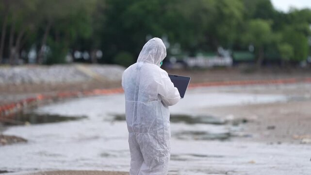 Person in protective coverall holding laptop at flooded site beside construction site, rear view during overcast weather, shallow water, tree and urban background during environmental field inspection
