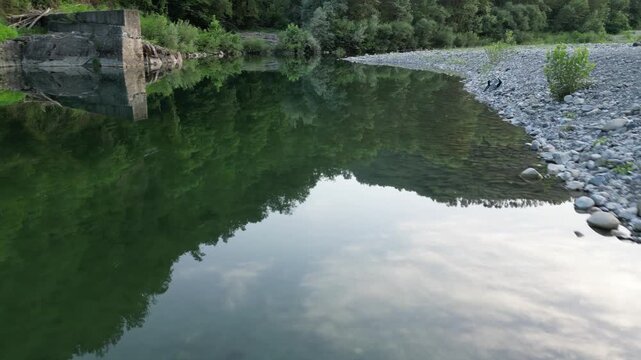 Aerial view of the clear waters of the Vara River flowing past a rocky riverbank with lush green trees in Province of La Spezia, Liguria, Italy.