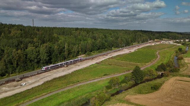 Drone view of a train traveling through tall forest along railway tracks with a broad open field extending into the distance and peaceful countryside surroundings