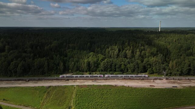 Aerial view of a passenger train traveling along railway tracks through a forest with an open green field stretching beside the railway under a clear daytime sky
