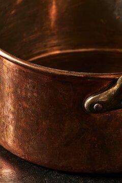 A close-up reveals the warm patina and hammered surface of an aged copper cooking pot, with its curved rim and dark metal handle catching soft light against a deep brown background