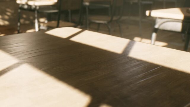 Sunlight streams through windows and casts shadows on a wooden table in a classroom. Empty chairs are visible in the background, indicating a time when students are not present.